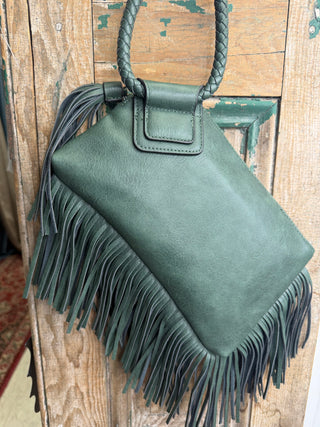 Olive green tassel fringe wristlet bag with braided ring handle and front flap, hanging on a rustic wood door