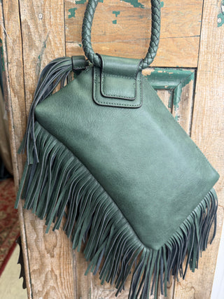 Close-up of olive green wristlet bag with long fringe trim and braided handle against weathered wood