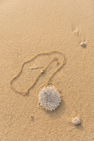 A gold chain necklace with a large, round, clear rhinestone-covered pendant, displayed on a rustic wood slice against a bright pink background.