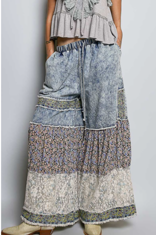 A person wearing loose, wide-leg patchwork pants with a blue-gray denim upper section and layered floral and textured fabric panels in beige, blue, and purple tones, paired with a gray ruffled top and beige boots against a plain light gray background.