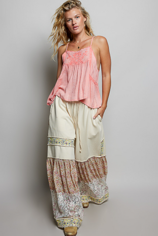 A woman modeling a bohemian-style outfit consisting of a light pink sleeveless camisole top and wide-leg cream pants with floral and patterned ruffle details, set against a plain light gray studio background.