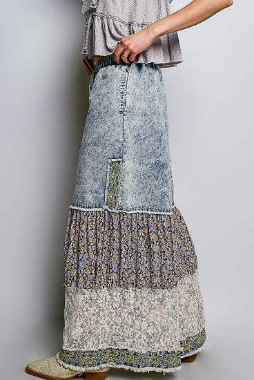 A long tiered denim maxi skirt with mixed fabric panels, including floral prints and lace, worn by a model. The skirt features a light blue acid-wash denim upper section and a multi-layered lower section with different patterns.