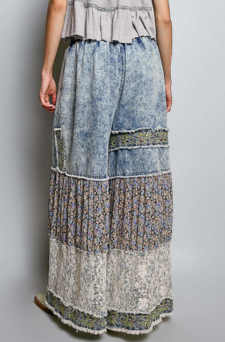 A person shown from the back wearing wide-leg patchwork pants with layered blue denim, floral print, and lace sections, against a plain light gray studio background.
