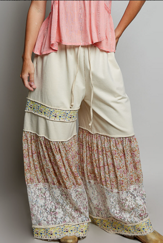 A person wearing loose, wide-leg cream pants with multicolored floral patterned lower sections and decorative embroidered bands, paired with a pink top against a plain light gray studio background.