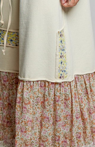 Close-up of a cream-colored knit top layered over a floral tiered skirt, featuring a vertical embroidered panel with blue, yellow, and green details.