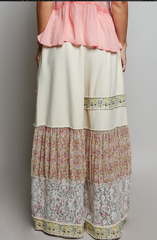 A person wearing a long, tiered maxi skirt with a solid cream upper section, a pink floral middle section, and a white floral lower section with decorative embroidered borders. The top is a light pink ruffled blouse, shown from the back against a neutral gray studio background.