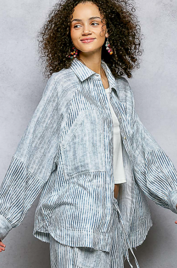 A woman with curly dark hair wearing a light blue and white vertically striped button-up shirt jacket with voluminous sleeves, styled over a white top and matching striped pants, set against a textured gray studio background.