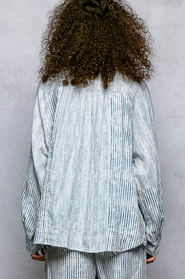 A person shown from the back wearing a loose, light blue and white vertically striped long-sleeve shirt and matching striped pants against a textured gray wall.