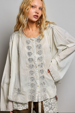 A woman wearing a loose, long-sleeve button-front blouse in a light warm off-white or cream fabric with gray floral embroidery down the front and on the sleeves, paired with patterned shorts, against a plain light gray studio background.