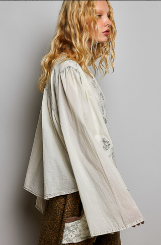 A woman shown in side profile wearing a loose, long-sleeved cream-colored blouse with delicate embroidery and a brown patterned skirt against a plain gray studio background.