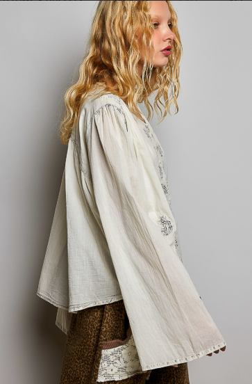A woman shown in side profile wearing a loose, long-sleeved cream-colored blouse with delicate embroidery and a brown patterned skirt against a plain gray studio background.