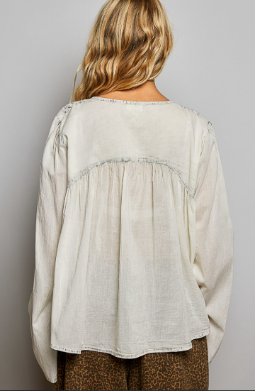 A woman viewed from the back wearing a loose, long-sleeve cream linen blouse with gathered detailing and frayed trim, paired with brown patterned pants, against a plain gray background.