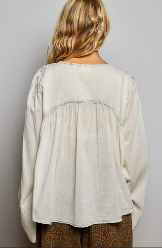 A woman viewed from the back wearing a loose, long-sleeve cream linen blouse with gathered detailing and frayed trim, paired with brown patterned pants, against a plain gray background.