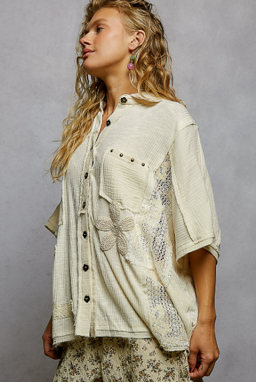 A woman wearing a cream-colored, textured short-sleeve button-up shirt with floral embroidery and lace applique, paired with matching patterned pants, set against a neutral gray studio background.