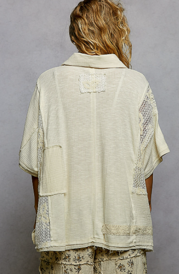 A person shown from the back wearing a cream-colored, textured short-sleeve shirt with lace panel details on the sides and a patch on the upper back, paired with patterned pants against a gray studio background.