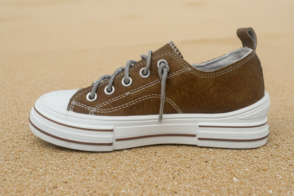 A brown suede low-top sneaker with a white rubber toe cap and sole, featuring silver rope laces threaded through metal eyelets, shown from a side profile on a sand  background.
