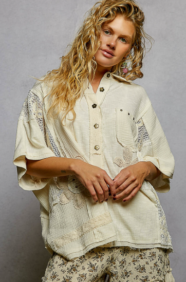 A woman wearing a cream-colored, bohemian-style short-sleeve button-up shirt with lace appliques and matching floral print pants, posed against a neutral gray studio background.