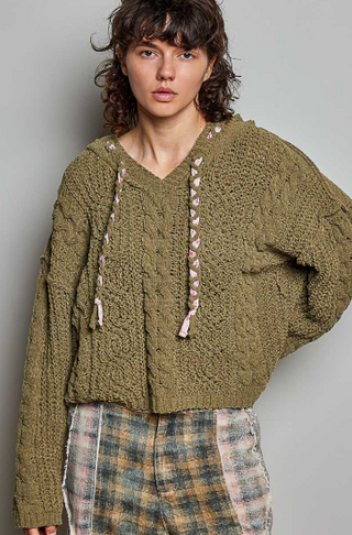 A model wearing a chunky brown cable-knit cropped hoodie sweater with pink and white braided drawstrings, paired with plaid pants, against a plain gray studio background.