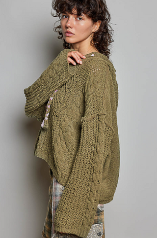 A woman wearing a chunky brown cable-knit hoodie sweater, shown from a side-back angle against a plain light gray studio background.