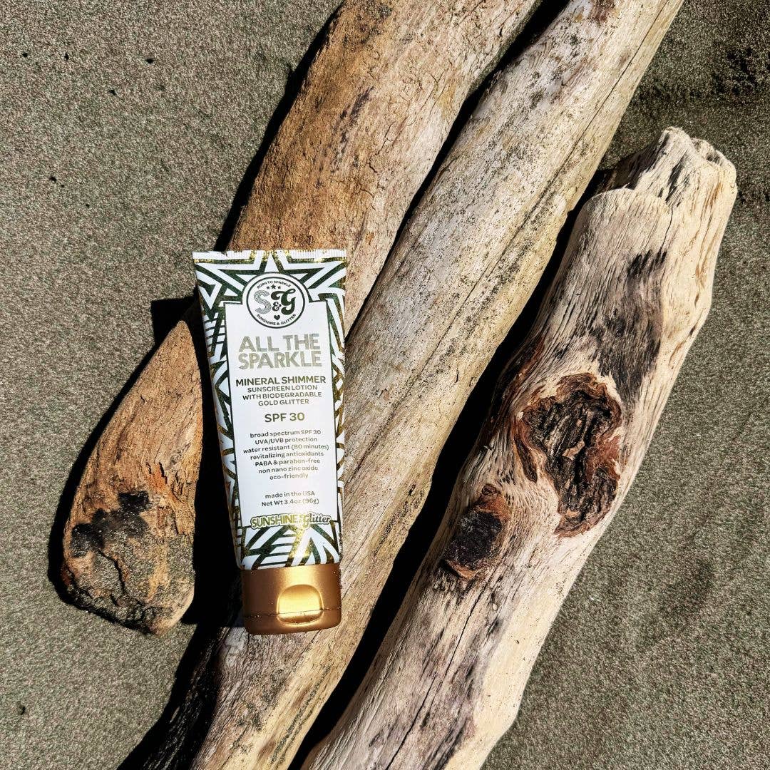 Tub of sunscreen with 'All the Sparkle' branding on a piece of driftwood on a sandy beach.