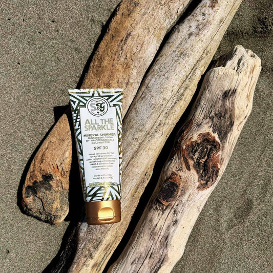 Tub of sunscreen with 'All the Sparkle' branding on a piece of driftwood on a sandy beach.