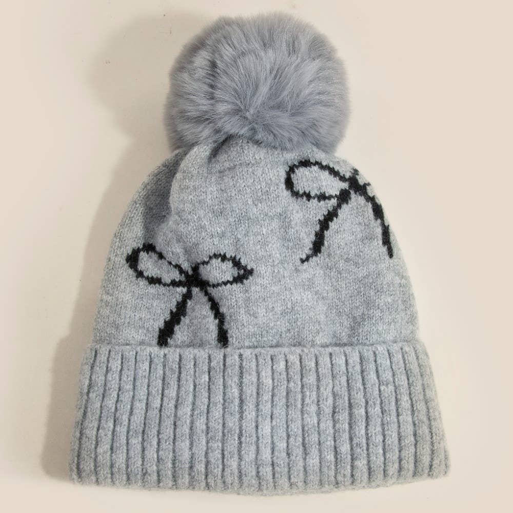 Gray knit beanie with pom-pom and black bow design on a beige background