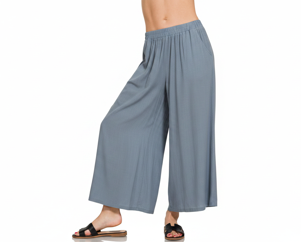 A person modeling wide-leg cropped pants in a dusty blue-gray color, shown from the waist down against a plain white background. The model is wearing black slide sandals