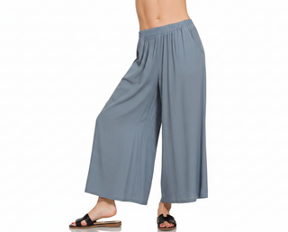 A person modeling wide-leg cropped pants in a dusty blue-gray color, shown from the waist down against a plain white background. The model is wearing black slide sandals