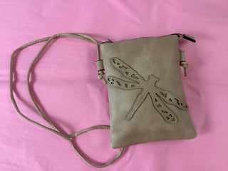 Tan perforated leather crossbody bag with silver studs, displayed on a wooden table in a retail setting with clothing racks and jewelry in the background