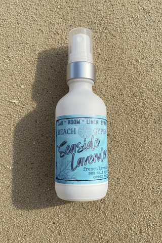 Glass bottle of Sea Witch coastal room spray resting on a sand-covered surface