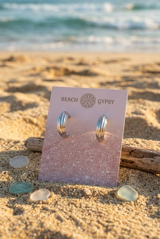 Silver-Tone Ridged Hoop Earrings with a thick, curved ridged design on a Beach Gypsy earring card, set in the sand by the ocean with driftwood and sea glass