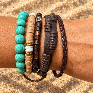A close-up of a person's wrist wearing multiple bracelets, including turquoise bead strands, tan and dark brown beaded bracelets, a braided brown leather bracelet, and a textured dark brown wrap bracelet.