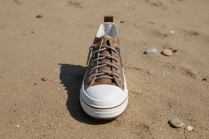 A single low-top sneaker shown from a front-facing overhead view on a sand background. The upper is primarily medium brown suede with a white rubber toe cap and white sole, and the laces appear sparkly silver.
